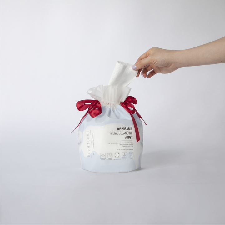 Hand pulling a disposable facial cleansing wipe from a stylish Dermalume packaging with red ribbon accents.